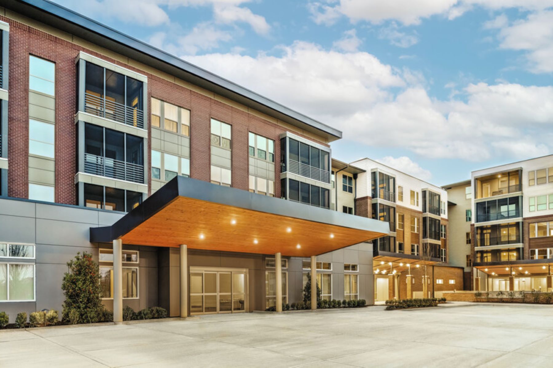 Modern senior living community with covered entryway, large windows, and multi-story brick and panel exterior under a blue sky.