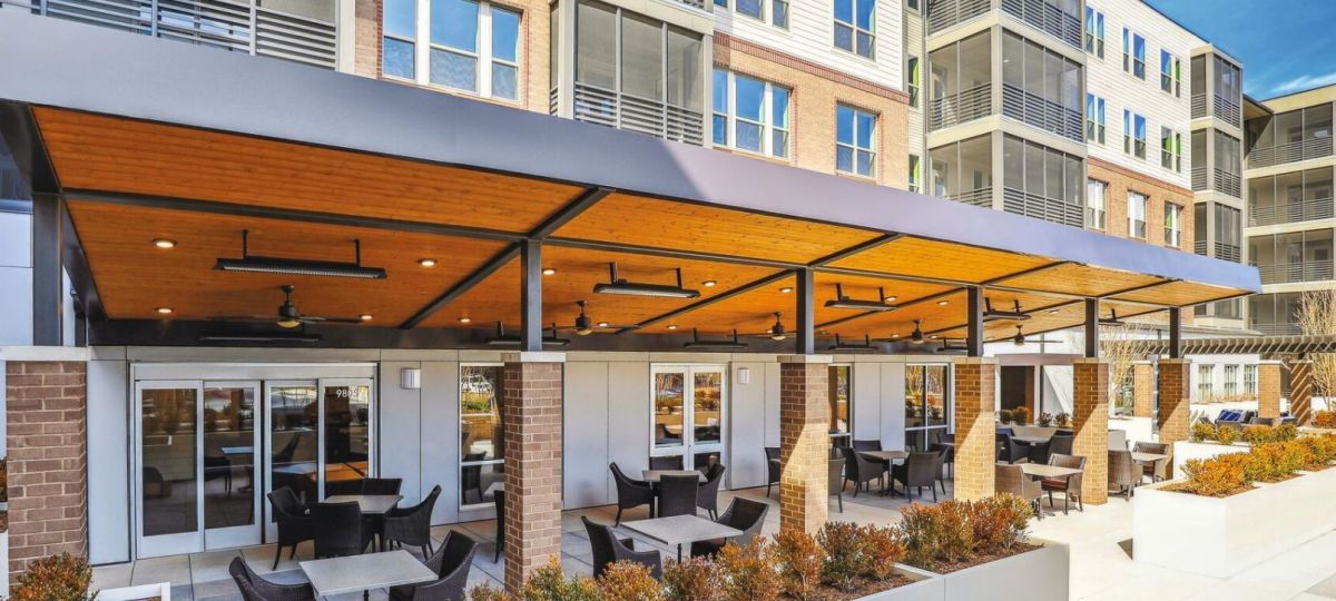 Covered outdoor patio with seating and ceiling fans at a modern apartment community courtyard, featuring wood-paneled canopy and landscaped planters.